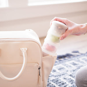 Woman puts littoes stacked food pots into side of diaper bag. Each food pot contains baby snack for a trip