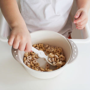 Toddler holds a spoon to scoop cereals against the wall of a bowl with side handles. 